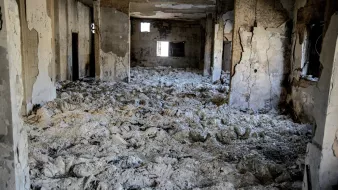 L'image montre l'intérieur d'un bâtiment en ruine, où les murs sont endommagés et décrépis. Le sol est recouvert d'un matériau blanc poussiéreux, probablement des débris ou des restes d'anciennes structures. Les fenêtres sont brisées ou manquantes, créant une atmosphère de désolation et d'abandon. La lumière pénètre à travers les ouvertures, mais l'espace parait sombre et sinistre.