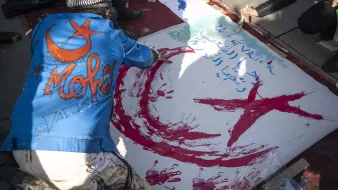 L'image montre une personne en train de peindre sur une grande toile au sol. La personne porte une veste bleu avec le mot "Moh" écrit dessus. On peut voir des éclaboussures de peinture rouge et verte, et un motif en forme de croissant de lune et d'étoile, qui sont des symboles souvent associés à certains pays. Des inscriptions sont également visibles sur la toile. L'environnement semble être celui d'une manifestation ou d'un évènement artistique.
