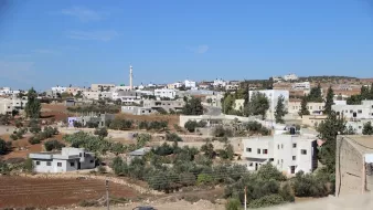 L'image montre un paysage urbain de type rural, avec des maisons de couleur claire disséminées sur une colline. On peut apercevoir des arbres et des champs, ainsi qu'un minaret se dressant dans le fond, ce qui pourrait indiquer la présence d'une mosquée. Le ciel est dégagé et d'un bleu vif, ce qui donne à la scène une atmosphère ensoleillée. Les infrastructures semblent simples et typiques des zones périphériques.