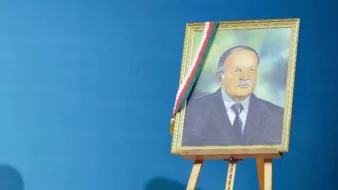 L'image montre un portrait encadré d'un homme, affiché sur un chevalet. Le fond est d'un bleu uniforme, créant un contraste avec le cadre doré du portrait. L'homme dans l'image a une expression sérieuse et porte un costume, accompagné d'un insigne ou d'un drapeau sur le cadre, possiblement représentant un symbole national ou officiel.