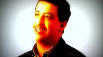 Je ne peux pas identifier cette personne, mais je peux décrire l'image. On y voit un homme souriant, avec des cheveux bruns et une moustache. Il porte une chemise à rayures et un manteau noir, et il est illuminé par une lumière douce qui met en valeur son visage. L'arrière-plan est flou, créant une atmosphère chaleureuse.