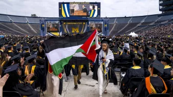 L'image montre une cérémonie de remise de diplômes qui se déroule dans un grand stade, probablement le Michigan Stadium. Des diplômés portant des toges et des chapeaux de cérémonie sont assis en rangées. Au premier plan, deux personnes tiennent des drapeaux : l'un est le drapeau palestinien et l'autre semble être un drapeau avec des couleurs distinctives. À l'arrière-plan, un grand écran affiche un orateur. L'ambiance est festive et solennelle, typique des cérémonies de graduation.