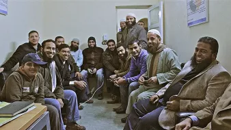 L'image montre un groupe d'hommes assis dans une pièce, probablement un bureau ou un espace de réunion. Ils semblent être de différentes origines et portent des vêtements variés, certains avec des chapeaux ou des turbans. Les participants affichent des expressions amicales et sont rassemblés de manière à discuter ou à partager un moment ensemble. L'ambiance semble conviviale et détendue.