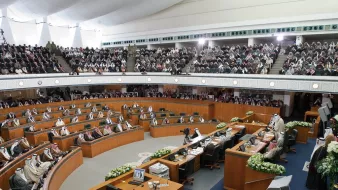 L'image montre une grande assemblée, probablement un parlement ou une salle de réunion officielle. On y voit des hommes en tenue traditionnelle, souvent vêtus de thobes et de keffiehs, assis à des bureaux en bois agencés en demi-cercle. La salle est bien aménagée, avec des décorations florales sur les tables et des écrans affichant des informations. Une grande audience est présente, ce qui suggère une session importante. L'ambiance semble solennelle et axée sur la discussion ou la prise de décision.