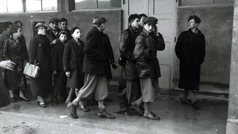 L'image montre un groupe de personnes, majoritairement des femmes et des enfants, marchant en direction d'un bâtiment. Ils portent des vêtements sombres et d'hiver. Leurs expressions semblent sérieuses et préoccupées. À l'entrée du bâtiment, une personne se tient à l'écart, observant le groupe avec une expression indéchiffrable. L'atmosphère de l'image est pesante et évoque un sentiment de tristesse ou d'incertitude. La scène semble avoir lieu dans un cadre urbain ou un lieu de rassemblement, avec des fenêtres en arrière-plan.