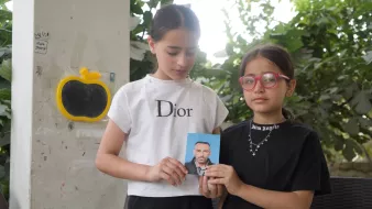 Sur l'image, on voit deux jeunes filles debout l'une à côté de l'autre. La fille de gauche porte un t-shirt blanc avec le logo "Dior" et tient en main une photo d'un homme. La fille de droite est vêtue d'un t-shirt noir avec des lunettes roses. Elles sont entourées par un arrière-plan verdoyant, avec des feuilles visibles autour d'elles.