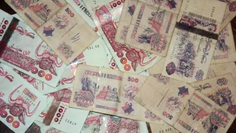 L'image montre un tas de billets de banque éparpillés, principalement de couleur beige avec des motifs et des textures variés. Certains billets sont de 500 unités, affichant des illustrations ou des inscriptions.