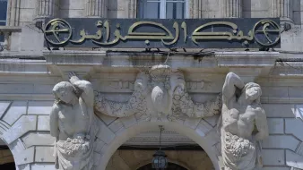 L'image montre une façade architecturale ornée d'une grande enseigne avec des inscriptions en arabe. Au-dessus de l'entrée, deux sculptures de figures humaines sont placées de part et d'autre, tenant chacune des éléments décoratifs comme des grappes de fruits. L'ensemble dégage un style classique, mettant en avant des détails sculptés sur la pierre et une embellissement artistique de l'architecture.