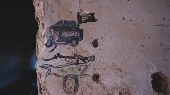 L'image montre un mur en béton décrépit où des graffitis sont visibles. On peut y voir une représentation d'un véhicule, probablement une voiture de police, accompagnée d'un drapeau. En dessous, il y a un dessin stylisé d'un squelette, surmonté d'une forme qui pourrait évoquer un traîneau ou un chariot. Le mur lui-même présente des traces d'usure et des fissures, ajoutant une dimension d'abandon à la scène.