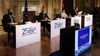 L'image montre une scène de conférence avec plusieurs intervenants assis derrière des panneaux portant le logo du processus de Barcelone, qui célèbre ses 25 ans. Les participants portent des masques, indiquant qu'ils suivent des mesures de santé, probablement en raison de la pandémie. À l'arrière-plan, on peut voir des drapeaux représentant l'Union européenne et l'Espagne, ainsi qu'un écran affichant des images, probablement liées à l'événement. L'environnement semble formel, suggérant une réunion diplomatique ou un sommet.