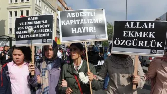 L'image montre une manifestation avec des personnes portant des pancartes. Les manifestants semblent exprimer des revendications contre la pression et l'oppression, en mettant en avant des messages en turc. Certaines femmes portent des foulards, suggérant un contexte culturel spécifique. On peut sentir une atmosphère de mobilisation collective.
