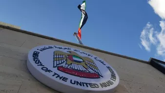L'image montre un bâtiment avec un panneau qui indique qu'il s'agit de l'ambassade des Émirats arabes unis. Au-dessus du panneau, on peut voir le drapeau des Émirats arabes unis flottant. Le ciel est clair et légèrement nuageux, ce qui donne une belle arrière-plan à la scène.