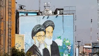 L'image montre une grande fresque murale représentant deux hommes avec des turbans noirs et des barbes blanches. Ils sont peints sur un mur d'un bâtiment, entourés de fleurs et de motifs décoratifs. En arrière-plan, on aperçoit des structures urbaines et un ciel légèrement nuageux. Cette fresque semble symboliser une figure ou un message important dans le contexte culturel ou historique de la région.