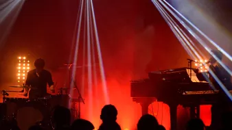 L'image montre une scène de concert avec deux musiciens. À gauche, un batteur joue de la batterie, tandis qu'à droite, un pianiste est assis devant un clavier. L'éclairage est dramatique, avec des faisceaux lumineux qui coupent à travers la fumée ambiante, créant une atmosphère dynamique. En arrière-plan, on devine une audience silhouette, profitant du spectacle. Les couleurs rouges et jaunes ajoutent à l'énergie de la performance.