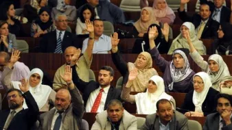 L'image montre un rassemblement dans un espace législatif, probablement au sein d'une assemblée ou d'un parlement. De nombreuses personnes sont présentes, avec une majorité levant la main, ce qui indique probablement un vote ou une approbation. Les participants semblent porter des tenues formelles, et certaines femmes portent le hijab. On observe une diversité de participants, tant en termes de genre que d'âge. L'environnement évoque une atmosphère de discussion et de prise de décision collective.