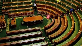 L'image montre l'intérieur d'une assemblée, probablement un parlement ou une salle de réunion officielle. On peut voir des rangées de sièges en bois oblongs disposés en arc de cercle, avec un grand bureau central ovale. Les bancs sont vides, ce qui donne une impression de calme. La décoration est sobre, avec des éléments en bois et un tapis vert. Deux personnes sont visibles, probablement en train de ranger ou d'entretenir l'espace. L'ambiance générale semble formelle et institutionnelle.