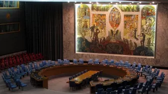 L'image montre l'intérieur d'une salle de réunion, probablement celle du Conseil de sécurité des Nations Unies. On y voit un grand panneau mural coloré représentant des scènes symboliques. La salle est circulaire avec une table ovale au centre, entourée de chaises disposées en arcs. Les chaises sont de différentes couleurs, majoritairement bleues et rouges. L'éclairage est doux, mettant en valeur les œuvres d'art sur le mur. L'atmosphère semble formelle et solennelle, propice aux discussions diplomatiques.