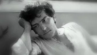 L'image montre un jeune homme pensif, appuyé sur sa main, avec une expression mélancolique. Il semble perdu dans ses pensées, ses cheveux sont légèrement ondulés et il porte une chemise à manches longues. L'arrière-plan est flou, créant une ambiance douce et introspective. La lumière est tamisée, ajoutant à l'atmosphère de réflexion.