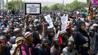L'image montre une grande foule de manifestants réunis, tenant des pancartes. Les personnes semblent exprimer leur mécontentement et leurs revendications. On peut voir une diversité de vêtements parmi les participants, allant des habits traditionnels à des vêtements plus modernes. L'atmosphère paraît énergique et engagée, avec des individus qui tentent de faire entendre leur voix. Des barrières sont présentes pour délimiter l'espace.