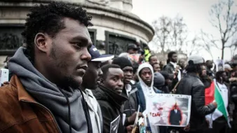 L'image montre un groupe de personnes rassemblées, probablement lors d'une manifestation ou d'un événement public. Au premier plan, un homme au port de tête pensif se distingue, entouré d'autres participants qui tiennent des pancartes et des photos. L'atmosphère semble engagée et solidaire, avec des affiches qui pourraient exprimer des revendications ou des messages de protestation. L'environnement urbain est visible en arrière-plan, suggérant que cet événement se déroule dans un espace public.