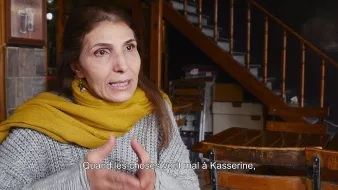 L'image montre une femme en train de parler, assise dans un environnement intérieur. Elle porte un vêtement gris avec un foulard jaune autour du cou. L'arrière-plan présente des escaliers en bois et des tables en bois, ce qui donne une ambiance chaleureuse et intime. Elle semble s'exprimer avec passion, évoquant probablement des enjeux importants liés à la ville de Kasserine.