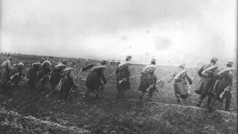 L'image montre un groupe de soldats marchant en formation sur un terrain accidenté. Ils portent des uniformes militaires, avec des chapeaux distinctifs, et semblent se déplacer dans une direction déterminée. Le paysage est marqué par un ciel nuageux et une ambiance sombre, suggérant peut-être un contexte de guerre ou de mouvement stratégique dans un environnement rural. Le sol est boueux, ce qui rend leur avance difficile. L'image est en noir et blanc, renforçant le caractère historique et poignant de la scène.