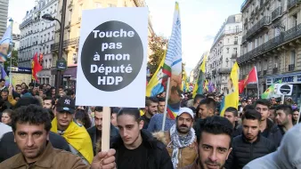 L'image montre une manifestation de foule dans une rue, où des personnes tiennent des pancartes et des drapeaux. Au premier plan, un manifestant porte une grande pancarte avec le message "Touche pas à mon Député HDP". L'ambiance semble être celle d'une protestation politique, impliquant probablement des questions liées au parti HDP (Parti démocratique des peuples) en Turquie. Les participants semblent engagés et déterminés.