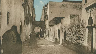 L'image représente une ruelle étroite et pavée, probablement dans une ville historique. Les murs des bâtiments sont en pierre, avec des textures vieilles et usées, suggérant un certain âge. On peut apercevoir des silhouettes de personnes marchant, certaines vêtues de longues robes traditionnelles. À l'arrière-plan, un arc architectural est visible, ajoutant une touche d'authenticité à l'environnement. Le ciel est dégagé, apportant une lumière douce à la scène. L'atmosphère semble paisible et évoque un lieu chargé d'histoire.