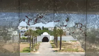 L'image montre une vue à travers un grand trou dans une vitre brisée. On peut voir un paysage urbain avec des palmiers et un édifice en arrière-plan, probablement une entrée ou un portail. Le ciel est dégagé et la scène semble être abandonnée, avec des graffiti visibles sur le verre. L'atmosphère de l'image pourrait évoquer un sentiment de désolation ou d'oubli.