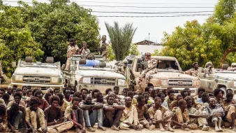 L'image montre un groupe de personnes assises au sol, semblant fatiguées et dans des vêtements usés, tandis qu'elles sont entourées de plusieurs véhicules tout-terrain. À l'arrière-plan, on peut voir des arbres et des plantes, suggérant un environnement rural. Certains individus semblent garder les véhicules, et l'atmosphère générale donne une impression de tension ou de détresse.