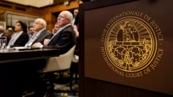 L'image montre une scène à la Cour internationale de justice. Au premier plan, on peut voir le symbole de la Cour, qui est un emblème distinctif. En arrière-plan, plusieurs personnes sont assises, probablement des membres du tribunal ou des délégués, attendant une session ou un discours. L'environnement semble formel, avec une décoration typique d'une institution judiciaire.