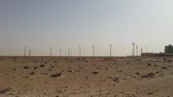 L'image montre un paysage désertique, caractérisé par un terrain aride avec des débris, probablement des déchets, éparpillés sur le sol. À l'arrière-plan, on peut voir plusieurs éoliennes qui s'élèvent vers le ciel clair. Des pylônes électriques sont également visibles, ajoutant une touche industrielle à ce paysage naturel. Le ciel est dégagé, ce qui accentue l'effet désertique de la scène.