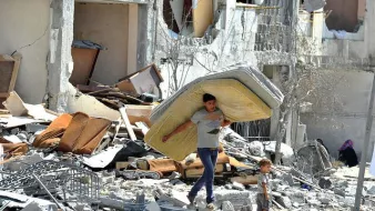 L'image montre un jeune homme portant un matelas sur son dos, marchant à travers des décombres. À côté de lui, un petit garçon l'observe. Le décor est celui d'un bâtiment en ruines, avec des meubles éparpillés et des murs endommagés, ce qui suggère une scène de destruction et de détresse. L'atmosphère est empreinte de tristesse et de désolation.