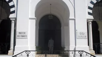 L'image montre l'entrée d'un bâtiment qui semble être le ministère des Finances, avec des inscriptions en français et en arabe. Le bâtiment est de style architectural classique, caractérisé par des murs blancs et des éléments décoratifs. On peut voir une grande porte d'entrée, entourée de fenêtres avec des grilles artistiques. À l'avant, une personne est visible, tournant le dos, s'approchant de l'entrée. Des plantes ornent l'espace autour des marches qui mènent à la porte.