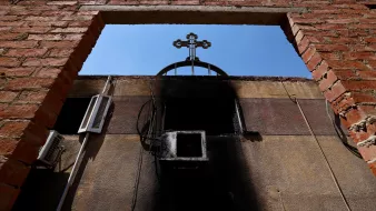 L'image montre un cadre en briques laissant entrevoir un ciel bleu, avec une croix noire au sommet d'une structure. On peut également voir des éléments d'électricité sur le mur, et une partie du mur semble avoir subi des dommages dus à un incendie, avec des marques noires. L'ensemble dégage une atmosphère de contraste entre la lumière et l'ombre, ainsi qu'une impression de désolation.
