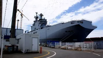 L'image montre un grand navire de guerre, probablement un porte-hélicoptères ou un navire amphibie, amarré dans un port. Le navire est de couleur grise et possède des structures sur le pont, suggérant qu'il est conçu pour des opérations militaires. En arrière-plan, on peut voir des grues et d'autres installations portuaires, tandis qu'à l'avant, il y a un petit bâtiment et des barrières de sécurité. Le ciel est partiellement nuageux, ajoutant une ambiance de chantier naval.