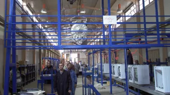 L'image montre un environnement d'atelier ou d'usine. On peut y voir des structures en métal, probablement des systèmes de rangement ou de support. Il y a des personnes travaillant dans cet espace, accompagnées d'équipements comme des appareils électroménagers, qui semblent être en phase de montage ou de préparation. L'éclairage est suffisant, et l'agencement des machines et des travailleurs suggère une ligne de production ou un processus d'assemblage. On note également des éléments rigides de la construction qui maintiennent l'espace organisé.