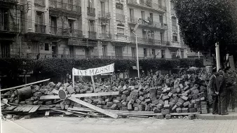 L'image montre une scène historique avec un amas de briques et de débris, suggérant une barricade ou un barrissement. Au premier plan, on peut voir des personnes rassemblées, probablement des manifestants ou des membres d'une foule, observant la scène. Un panneau est visible, ce qui pourrait indiquer le message ou le motif de la manifestation. À l'arrière-plan, des bâtiments avec des balcons sont présents, typiques d'une architecture urbaine. L'atmosphère semble tendue, reflétant un moment de contestation ou de protestation.