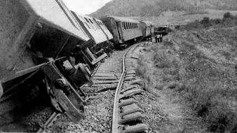 L'image montre un accident de train. Plusieurs wagons sont déraillés, penchés sur le côté, tandis que les rails semblent déformés. En arrière-plan, on aperçoit des montagnes et des personnes qui semblent inspecter les lieux. L'atmosphère est celle d'un événement tragique, évoquant une situation d'urgence.