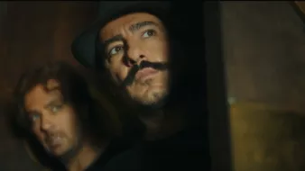 L'image montre deux hommes. L'un d'eux a une moustache prononcée et porte un chapeau, tandis que l'autre a les cheveux bouclés et semble inquiet. Ils regardent tous les deux vers la droite, comme s'ils étaient en train d'espionner ou de guetter quelque chose. L'éclairage est sombre, ce qui ajoute une atmosphère tendue et mystérieuse à la scène.