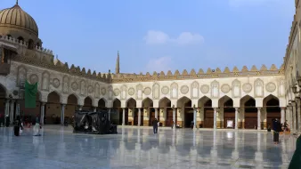 L'image montre une cour spacieuse d'un bâtiment historique, probablement un lieu de culte ou une mosquée. Le sol est en marbre poli, reflétant la lumière du ciel. On peut observer des arches élégantes et de nombreuses colonnes tout autour, ornées de motifs architecturaux détaillés. Au fond, on aperçoit un dôme et des minarets qui s'élèvent dans le ciel. La scène est calme, avec quelques personnes se déplaçant dans l'espace. Le ciel est clair, parsemé de nuages légers.