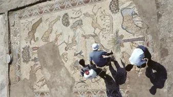 L'image montre une vue aérienne d'un sol en mosaïque impressionnant, découvert lors de fouilles archéologiques. Cette mosaïque représente une variété d'animaux, tels que des lions et des cerfs, ainsi que des motifs floraux et géométriques. On peut voir deux personnes travaillant à proximité, probablement en train d'examiner ou de restaurer la mosaïque. La terre autour de la mosaïque est dénudée, suggérant un site archéologique en plein exploration. Les ombres des personnes ajoutent une dimension intéressante à la scène.