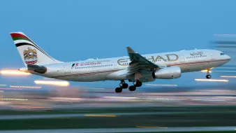 L'image montre un avion de la compagnie aérienne Etihad Airways en phase de décollage ou d'atterrissage. L'avion est partiellement flou, ce qui suggère une vitesse élevée. Le ciel apparaît dans une teinte bleue, tandis que des lumières brillantes en arrière-plan ajoutent une ambiance dynamique. L'appareil affiche la décoration et le logo caractéristiques de la compagnie.