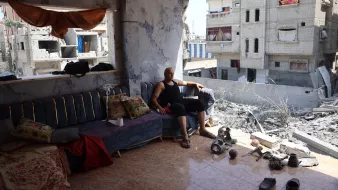 L'image montre un homme assis sur un canapé dans une pièce endommagée. Il semble se reposer, vêtu d'un débardeur noir et d'un pantalon court et noir. La pièce est en mauvais état, avec des murs endommagés et des débris au sol. À l'extérieur de la pièce, on aperçoit des bâtiments gravement endommagés, suggérant qu'il s'agit d'une zone touchée par un conflit ou une catastrophe. Le mobilier à l'intérieur, comme le canapé et certains coussins, semble également en désordre. L'atmosphère générale est celle de la dévastation et de la récupération après un événement destructeur.