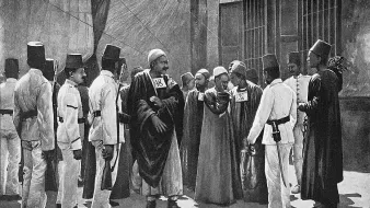 L'image présente un groupe de personnes en tenue traditionnelle. Au centre, un homme en robe longue semble diriger la scène, tandis que d'autres, habillés en uniformes ou en costumes distinctifs, l'entourent. Certains des individus portent des chapeaux spécifiques, comme des fez. Ils semblent engagés dans une conversation ou une activité, suggérant une interaction communautaire ou une réunion formelle. L'arrière-plan indique un environnement urbain, peut-être une scène historique. La composition dégage une atmosphère d'autorité et de respect.