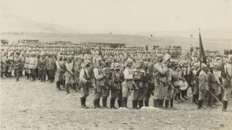 L'image montre une grande formation de soldats en uniforme, probablement d'un conflit militaire historique. Ils sont alignés en rangées serrées sur un terrain désertique ou peu végétalisé. Certains soldats portent des armes et semblent être en position de marche ou de formation. En arrière-plan, on peut voir d'autres soldats formant également des rangées, ce qui donne une impression de mobilisation de grandes troupes. L'atmosphère est sérieuse et solennelle, évoquant le contexte militaire de l'époque.