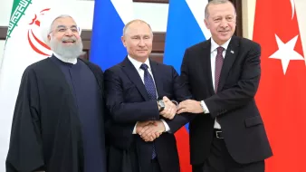 L'image montre trois dirigeants se tenant côte à côte. À gauche, un homme portant une robe sombre et une barbe grise, au centre un homme en costume sombre avec une cravate, et à droite, un homme également en costume qui semble sourire. Ils se tiennent par les bras, affichant une expression amicale, devant des drapeaux représentant leurs pays respectifs. L'environnement semble être une salle de réunion ou une conférence.