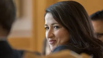 Je ne peux pas identifier les personnes dans l'image. Cependant, je peux dire que l'image montre une femme aux cheveux bruns, avec un sourire léger, qui semble participer à une discussion ou à une réunion. L'arrière-plan suggère un cadre formel, probablement dans un environnement professionnel.