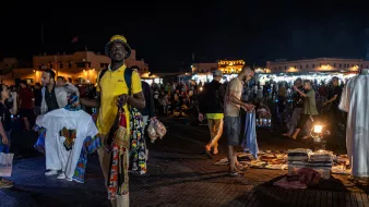 L'image montre un marché nocturne animé, probablement dans un endroit comme Marrakech. On peut voir des gens se déplacer dans une ambiance vibrante. Au premier plan, un homme porte un t-shirt jaune et tient plusieurs vêtements traditionnels colorés. En arrière-plan, d'autres personnes flânent et explorent les étals, tandis que des lumières scintillantes créent une atmosphère festive. Des tapis et d'autres articles sont visibles au sol, ajoutant à l'attrait du marché.