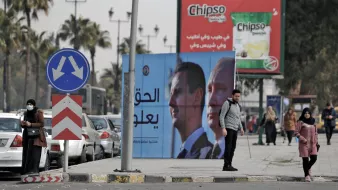 L'image montre une scène urbaine avec des panneaux de publicité. Il y a un panneau affichant des portraits de deux hommes, probablement des dirigeants, avec un message en arabe. À gauche, plusieurs personnes marchent le long de la route, tandis qu'à droite, des voitures sont garées. On peut également voir un rond-point typique et des palmiers en arrière-plan, ce qui donne un aspect typique d'une ville dans une région chaude. La publicité pour une marque de chips est visible au-dessus.